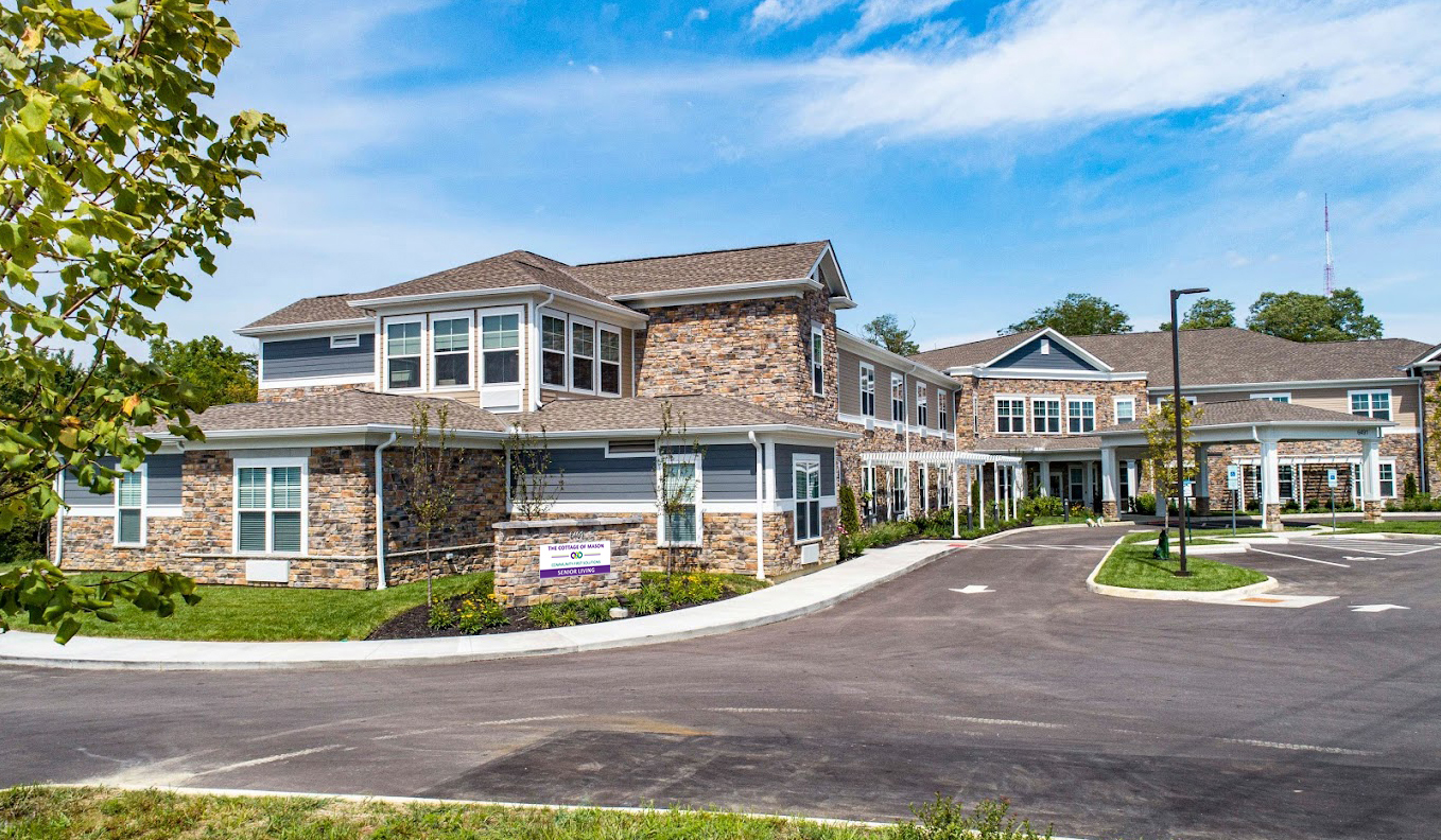 Exterior_TCOM-Sign_Mockup_0625_Web Two-story stone and siding senior living facility with covered entrance, multiple wings, landscaped grounds, and paved driveway on sunny day